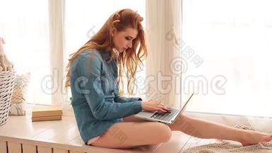 漂亮的红头发<strong>女士</strong>，马尾辫坐在宽阔的窗台上。 使<strong>用笔记本电脑</strong>和梦想。